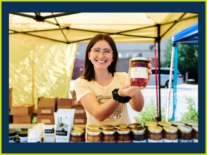 A woman stands at a farmers market, proudly holding a jar of homemade jam, showcasing local produce and craftsmanship.