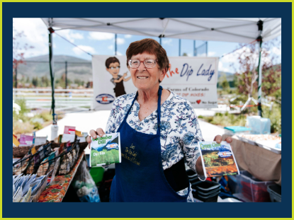A woman at a farmers market holds dip packet, immersed in the colorful surroundings of fresh fruits and vegetables.