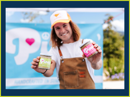 A man wearing an apron proudly displays two jars of peanut butter, emphasizing his passion for homemade treats.