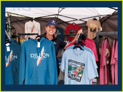A woman stands in front of a tent displaying a colorful assortment of t-shirts for sale.