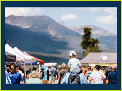 A father carries his son on his shoulders, enjoying a lively outdoor market filled with colorful stalls and people.