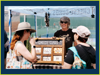 A woman sells various cheeses at a vibrant outdoor market, surrounded by fresh produce and eager customers.