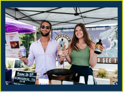 Two friends stand by a table, engaging in conversation with a bottle of tea placed on it.