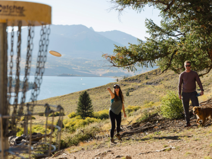 A man and a woman play frisbee golf on a grassy hillside under a clear blue sky
