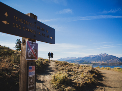 Two hikers walk along a scenic trail, passing a sign that provides information about the area