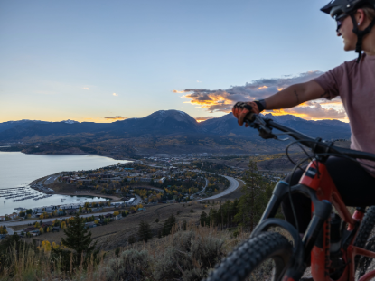 A cyclist on a mountain bike enjoys a scenic view of a tranquil lake framed by hills and trees.
