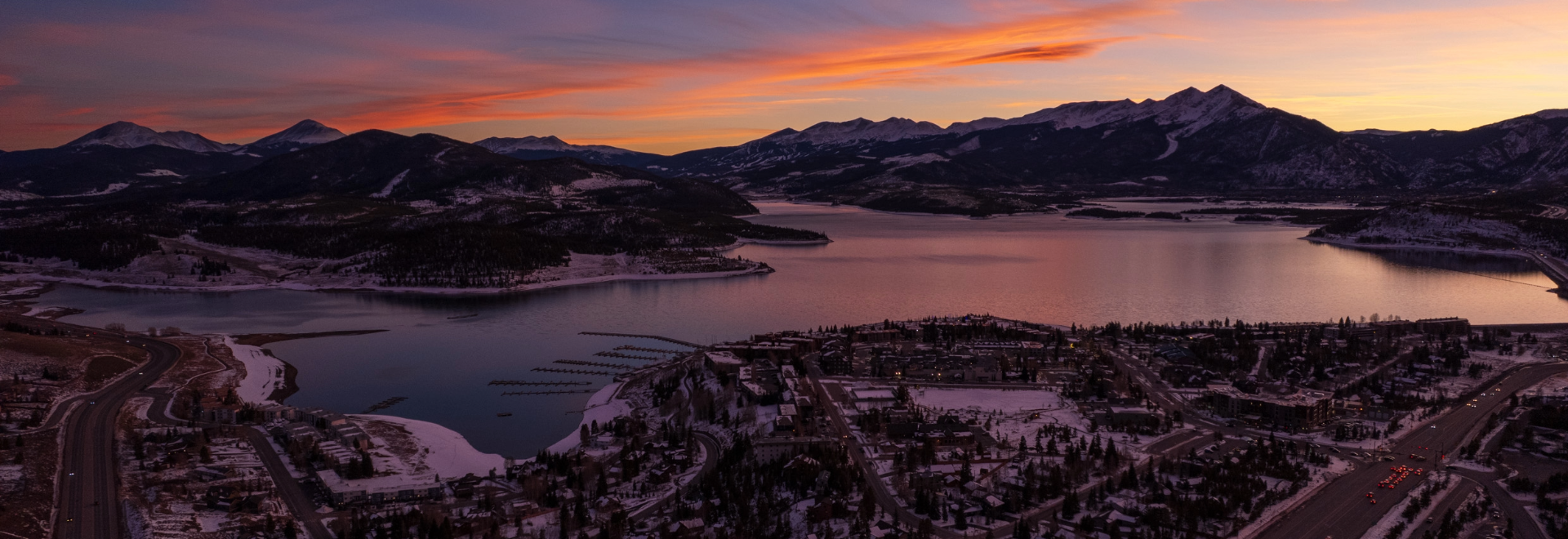 Sunset casts warm hues over mountains and a lake, framing a tranquil town at the water's edge.