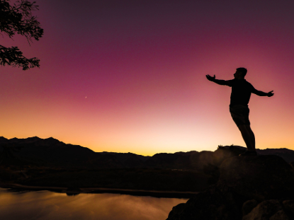 Man standing triumphantly on a mountain peak during a vibrant sunset, with colorful skies in the background