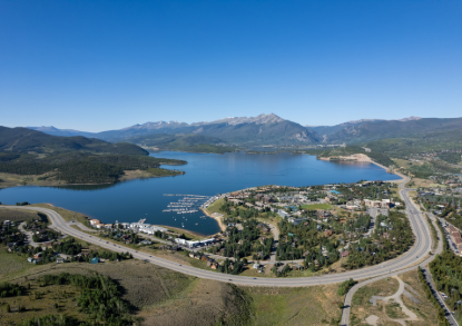 Aerial view of Dillon, Colorado, with Dillon Reservoir, marina, surrounding neighborhoods, and mountain peaks under a clear blue sky.