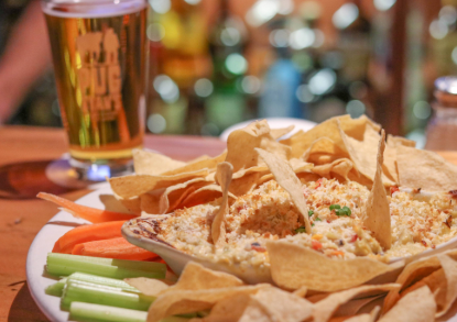 Basket of tortilla chips with a creamy dip, celery and carrot sticks, and a pint of beer on a wooden table in a bar setting.