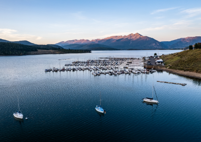 Boats are docked at a marina surrounded by mountains, reflecting a serene landscape of water and peaks