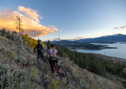 Two cyclists navigate a mountain trail on their bikes, surrounded by lush greenery and rocky terrain