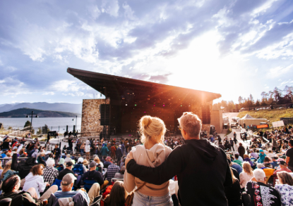 A large crowd of people enjoying an outdoor concert, with colorful lights and a stage in the background.